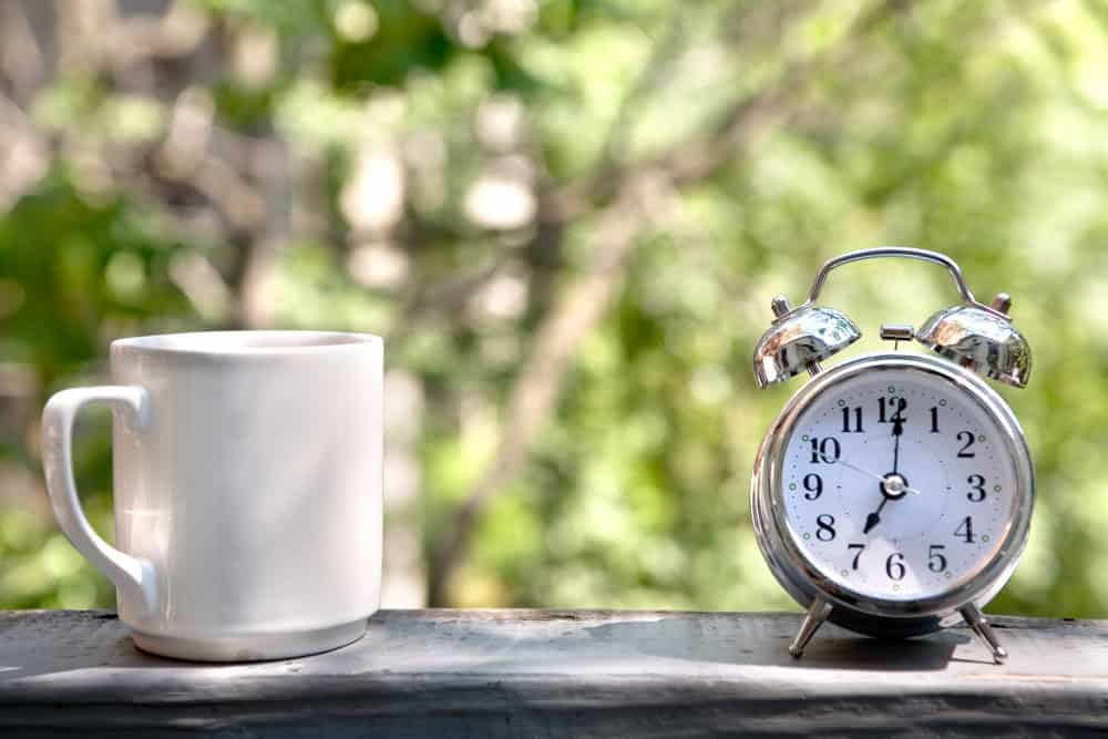 A white mug and a silver alarm clock on a ledge with a blurred outdoor background