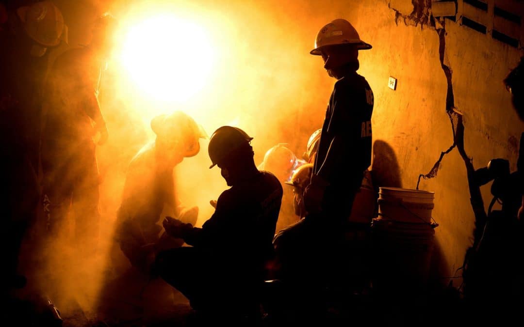 Silhouetted rescue workers in hard hats performing emergency response in a dusty environment.