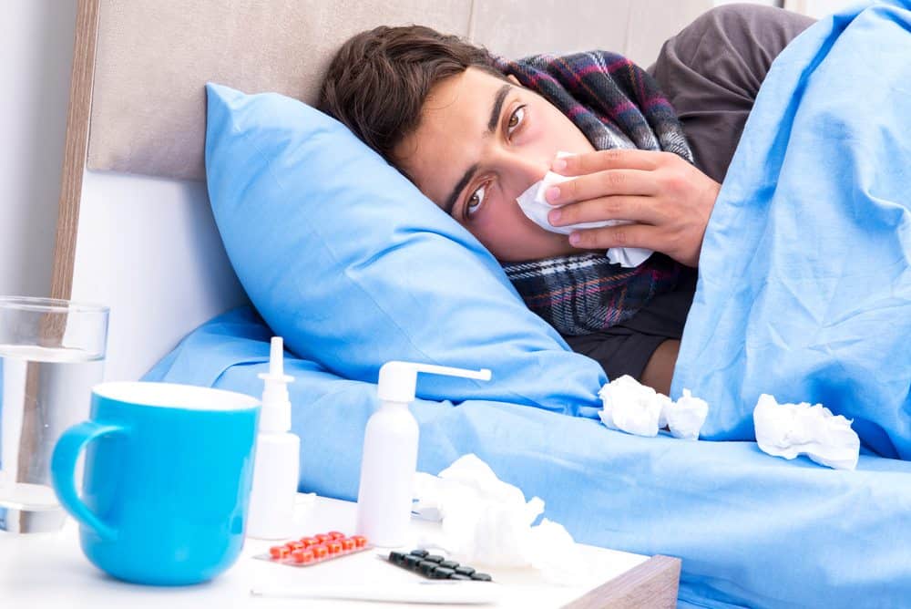 Sick person lying in bed with tissues, medication, and a cup nearby, showing illness recovery.