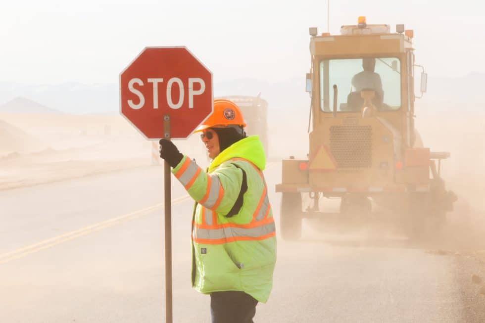 Road worker holding a stop sign with heavy machinery in a dusty construction area.