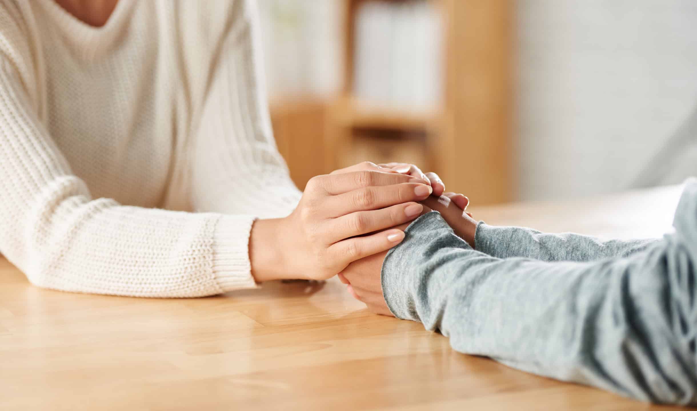 Close-up of two people holding hands in a gesture of comfort and support at a table.