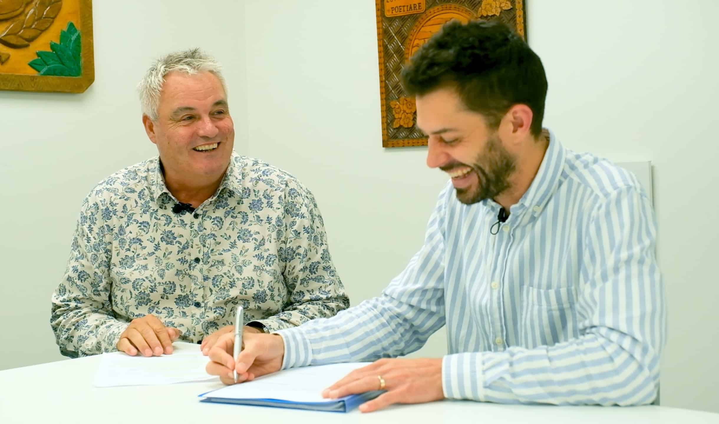 Two men smiling and signing documents during a meeting at an office.